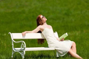 Red hair woman relaxing with book on white bench in a meadow; shallow DOF clipart