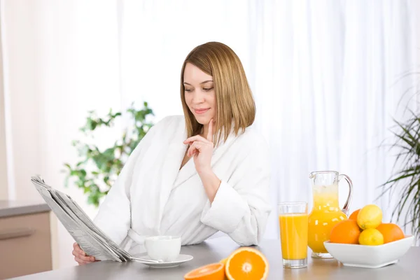 コーヒー 新鮮なオレンジ ジュース付きのキッチンで女性の読書新聞の笑みを浮かべて — ストック写真