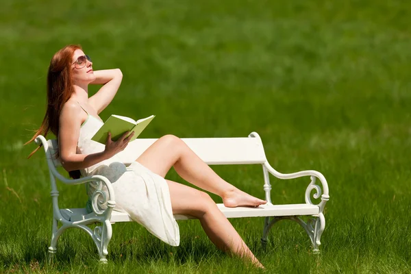 在绿色的草地上 浅景深的白色长椅上红头发的女人阅读书 — 图库照片