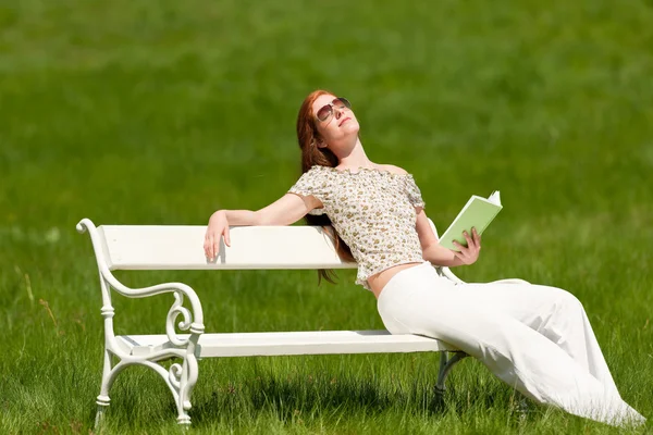 赤い髪の女性 草原で白いベンチで日光浴を楽しんでいます 浅い被写し界深度 — ストック写真