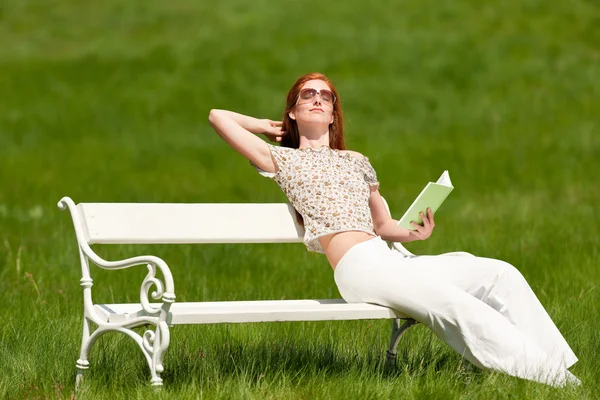 红头发的女人坐在一起坐在一片草地 白色长椅上的书浅景深 — 图库照片