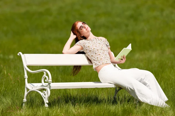 赤い髪の女性 草原で白いベンチに本を読んで浅い被写し界深度 — ストック写真