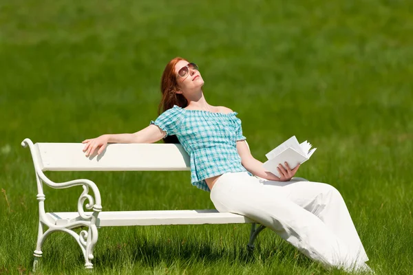 緑の牧草地 浅い被写し界深度で白いベンチで日光浴を楽しむ本赤髪の女性 — ストック写真