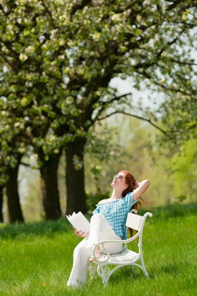 红头发女人坐在绿色的草地上 浅景深的白色长椅 — 图库照片