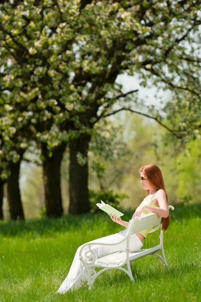 赤い髪の女性 草原で白いベンチに緑の本を読んで浅い被写し界深度 — ストック写真