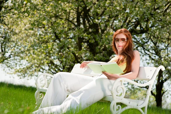 緑の牧草地 浅い被写し界深度で白いベンチの赤髪の女性読書 — ストック写真