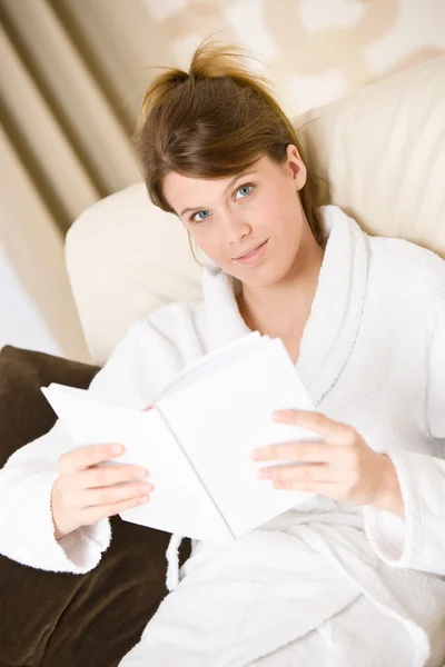 年轻女子读本书上沙发放宽在休息室穿着浴袍 — 图库照片