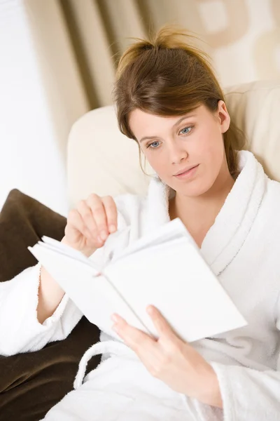 年轻女子读本书坐在沙发放宽在休息室穿着浴袍 — 图库照片
