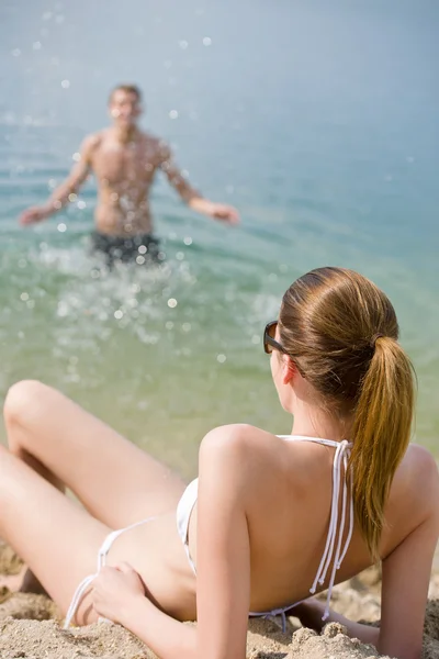 ビキニで日光浴海ビーチに男水のしぶきをバック グラウンドでの女性 — ストック写真