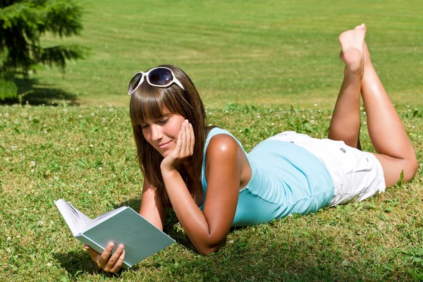 Χαμογελώντας νεαρή γυναίκα ξαπλωμένος στο γρασίδι με βιβλίο — Φωτογραφία Αρχείου
