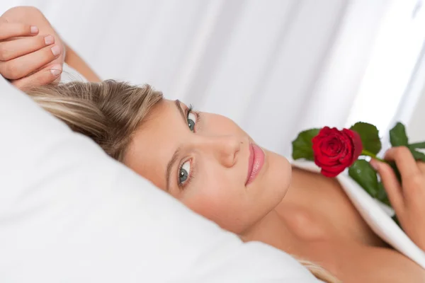White lounge - Beautiful woman lying in bed holding red rose — Stock Photo, Image