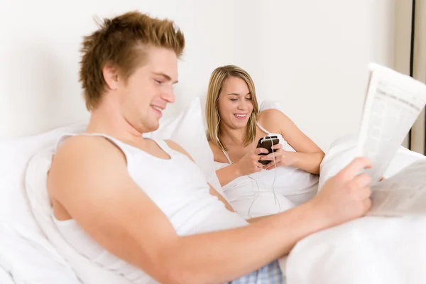 年轻夫妇精美的白色床上听听音乐 读报纸 — 图库照片