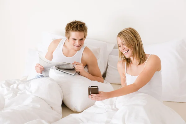 年轻夫妇精美的白色床上听听音乐 读报纸 — 图库照片
