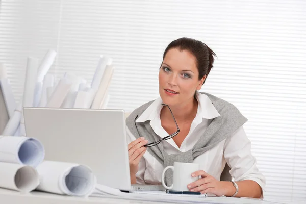微笑与便携式计算机和咖啡坐在办公室的女性建筑师 — 图库照片