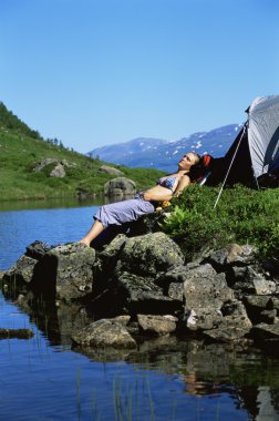 Young woman sunbathing on rocks next to lake clipart