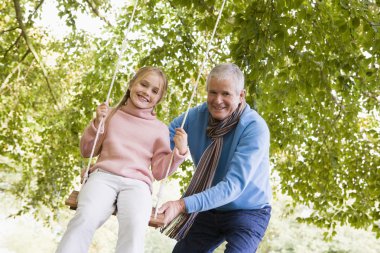Grandfather puching granddaughter on garden swing clipart