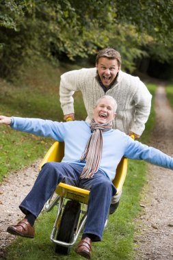 Grown up son pushing father in wheelbarrow along autumn path clipart