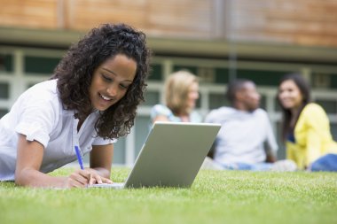Young woman using laptop on campus lawn, with other students rel clipart