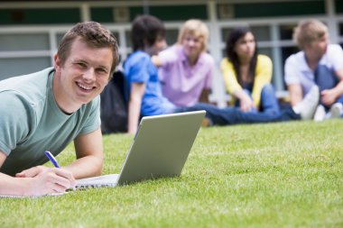 Young man using laptop on campus lawn, with other students relax clipart