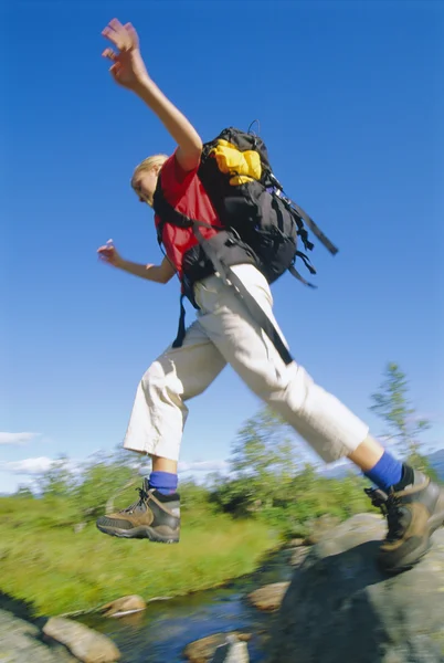 stock image Young woman wearing backpack leaping across river