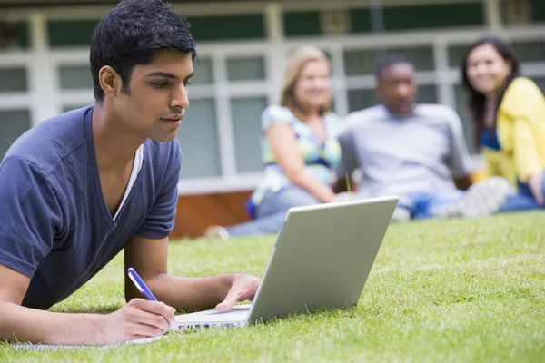 Jonge man met behulp van laptop op campus gazon, met andere studenten ontspannen — Stockfoto