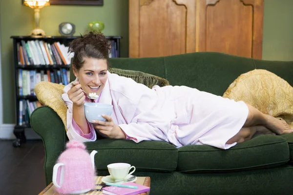 En ung kvinna liggande på hennes soffa äter spannmål — Stockfoto