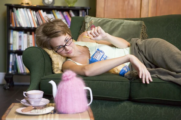 一个年轻的女人躺在她沙发上吃 — 图库照片