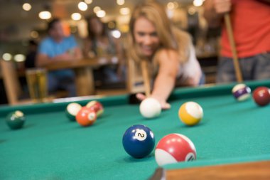 Young woman playing pool in a bar (focus on pool table) clipart