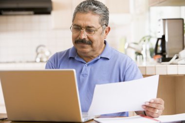 Man in kitchen with laptop and paperwork clipart