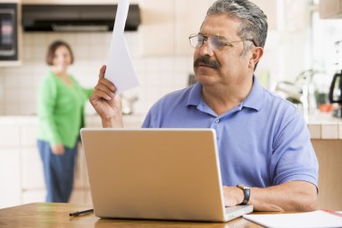 Man in kitchen with laptop and paperwork with woman in backgroun clipart