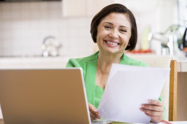 Woman in kitchen with laptop and paperwork smiling clipart