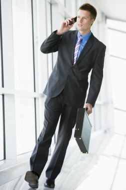 Businessman walking in corridor using cellular phone clipart