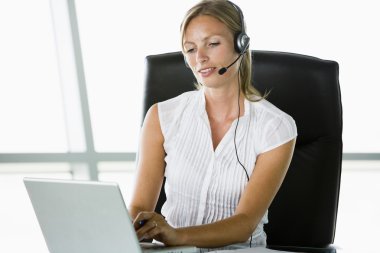 Businesswoman sitting in office wearing headset using laptop clipart