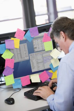 Businessman in office typing at computer with notes on it clipart