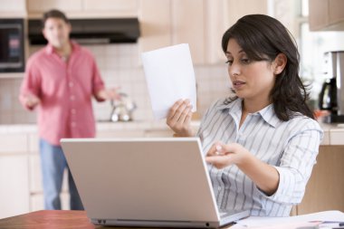 Woman in kitchen with paperwork using laptop with man in backgro clipart