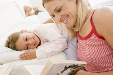 Woman reading book to young girl in bed smiling clipart