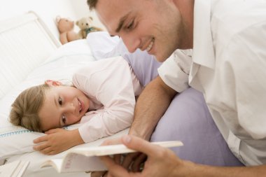 Man reading book to young girl in bed smiling clipart