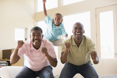 Two men and young boy in living room cheering and smiling clipart