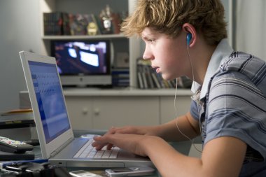 Young boy in bedroom using laptop and listening to MP3 player clipart