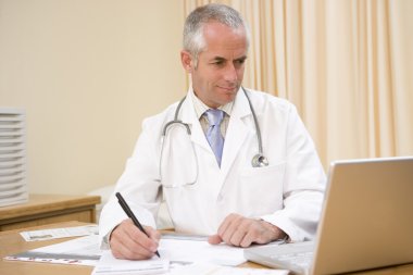 Doctor using laptop and writing in doctor's office clipart