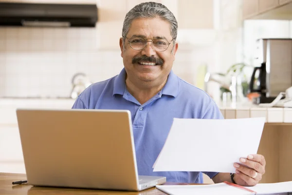 Человек на кухне с ноутбуком и бумажной работой улыбается — стоковое фото