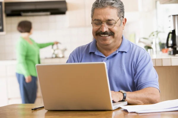 Man i köket med laptop leende med kvinna i bakgrunden — Stockfoto