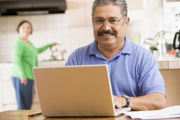 Homem na cozinha com laptop sorrindo com mulher no fundo — Fotografia de Stock