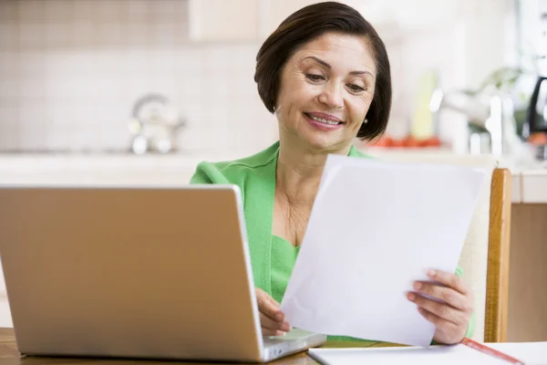 Mujer en cocina con laptop y papeleo sonriendo —  Fotos de Stock