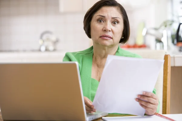 Femme dans la cuisine avec ordinateur portable et la paperasserie à l'air inquiet — Photo
