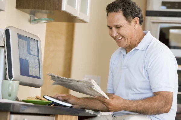 Homem na cozinha com computador e jornal sorrindo — Fotografia de Stock