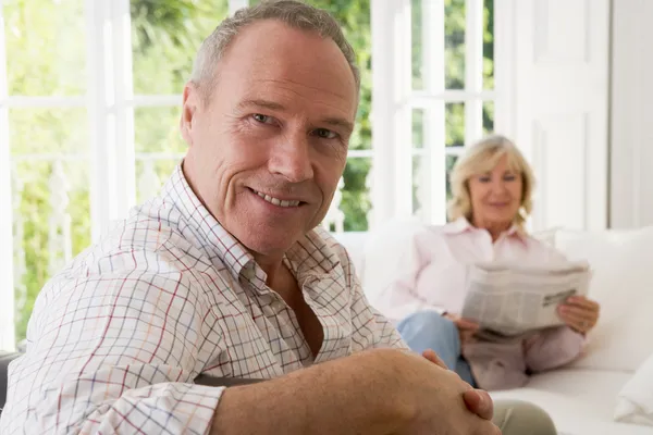 Man in living room smiling with woman in background reading news — Stock Photo, Image