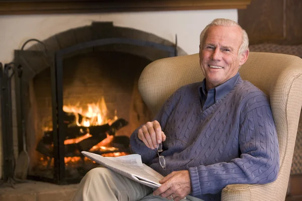 Hombre sentado en la sala de estar junto a la chimenea con periódico sonriendo —  Fotos de Stock