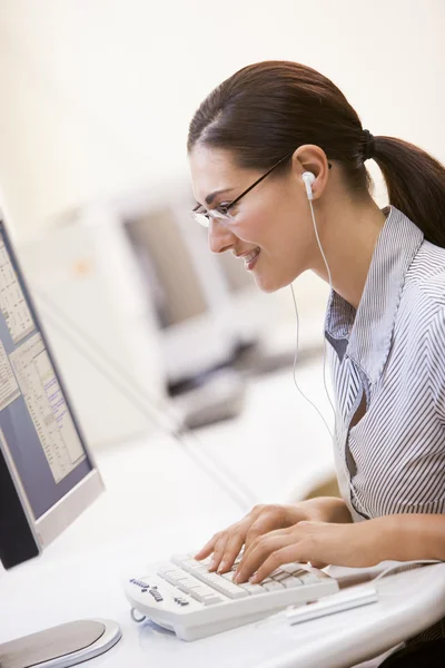 Mujer en la sala de computadoras escuchando MP3 Player mientras escribe y —  Fotos de Stock