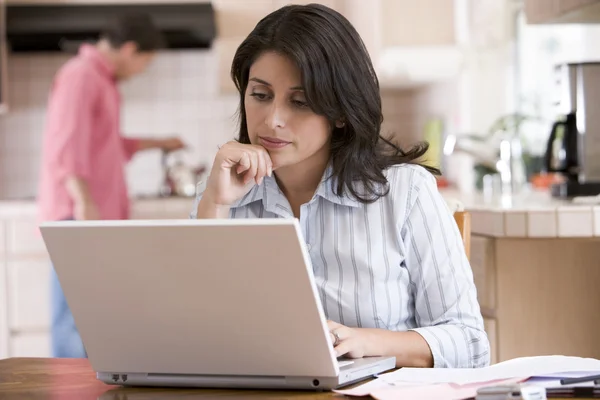 Женщина на кухне с бумажной работой с помощью ноутбука с мужчиной в backgro — стоковое фото
